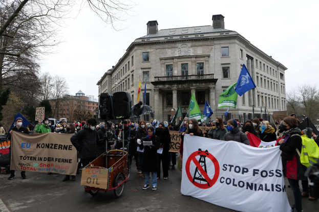 Große Gruppe von Menschen bei einer Demonstration gegen fossile Brennstoffe, die Schilder und Fahnen tragen, mit einem Fahrzeug im Vordergrund und Gebäuden, Bäumen und einem klaren Himmel im Hintergrund.
