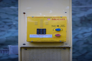 Ein gelbes Nottelefon an einer Gebäudeseite angebracht, mit Tasten und Text für Notrufe, vor einer Wand.