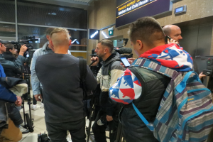 Eine Gruppe von Menschen, einige mit Kameras und Handys, andere mit Taschen, steht in einem Flughafen-Pressebereich mit einem Textschild und Lichtern dahinter.