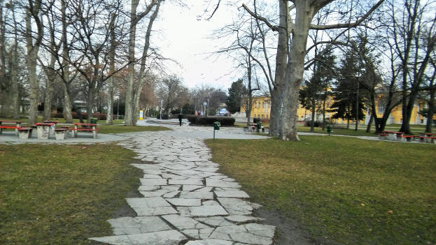 Ein Park mit einem gewundenen Steinweg, grünem Gras, Bäumen, Bänken, Tischen, Gebäuden und einem klaren blauen Himmel.