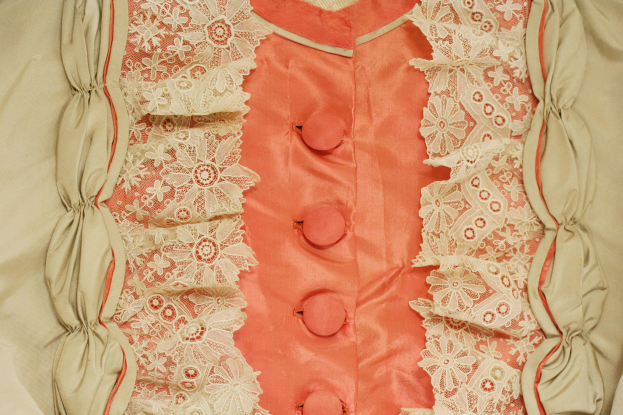 Nahaufnahme eines rosafarbenen und cremefarbenen Kleides mit weißer Spitzenborte und Knöpfen vorne.