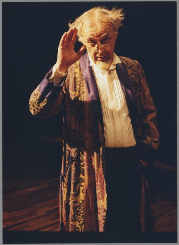 Ein Mann in einem langen Gewand und Brille steht auf einer Bühne mit gefalteten Händen vor einem Publikum gegen einen dunklen Hintergrund.