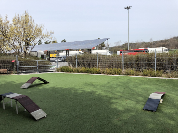 Hundepark mit Kunstgras, Rampen, Einzäunung, Grünflächen, Fahrzeugen, Schuppen, Pfosten und einer Laterne unter einem sichtbaren Himmel.