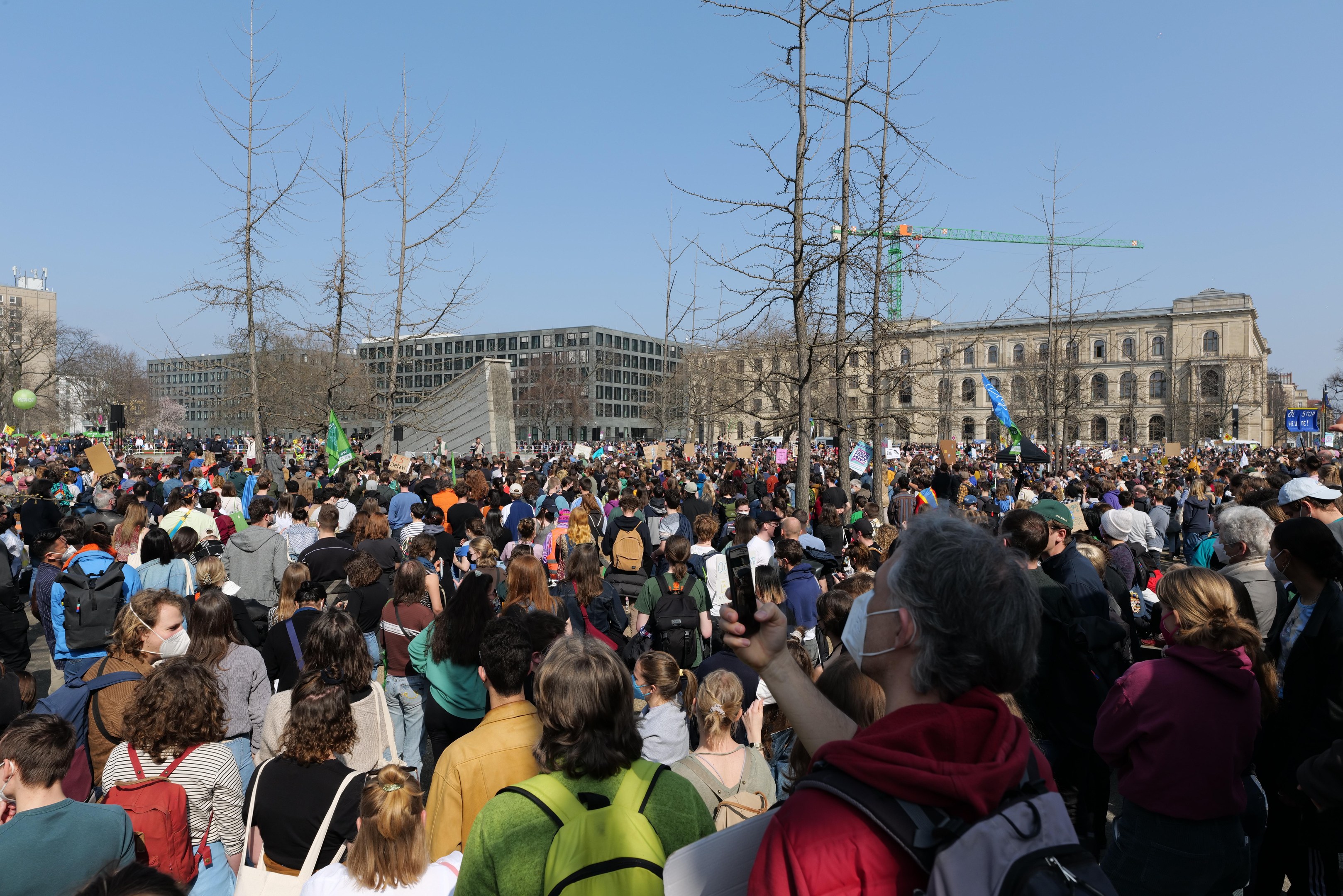 Eine große Menge Menschen steht vor einem Gebäude mit Fenstern, Bäumen und einem klaren Himmel, viele halten Schilder und tragen Taschen, was auf eine Klimaprotestveranstaltung hindeutet.