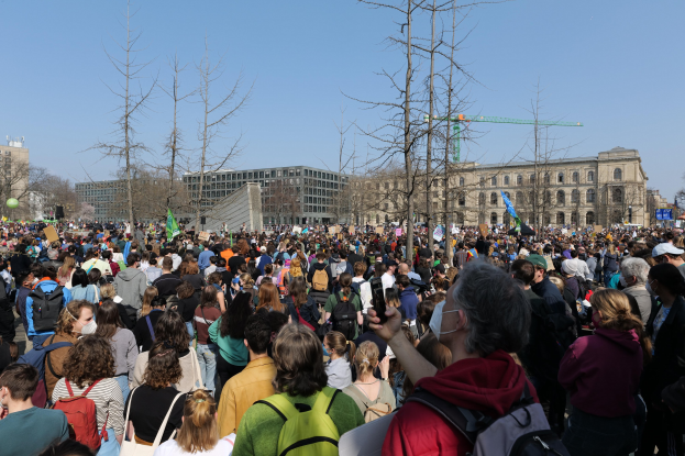 Eine große Menge Menschen steht vor einem Gebäude mit Fenstern, Bäumen und einem klaren Himmel, viele halten Schilder und tragen Taschen, was auf eine Klimaprotestveranstaltung hindeutet.