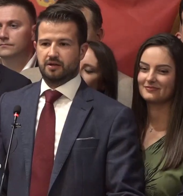 Ein Mann im Anzug spricht an einem Rednerpult mit einem Mikrofon zu einer Menge hinter einem Banner, wahrscheinlich diskutiert er ein Regierungsverbot von Social Media.