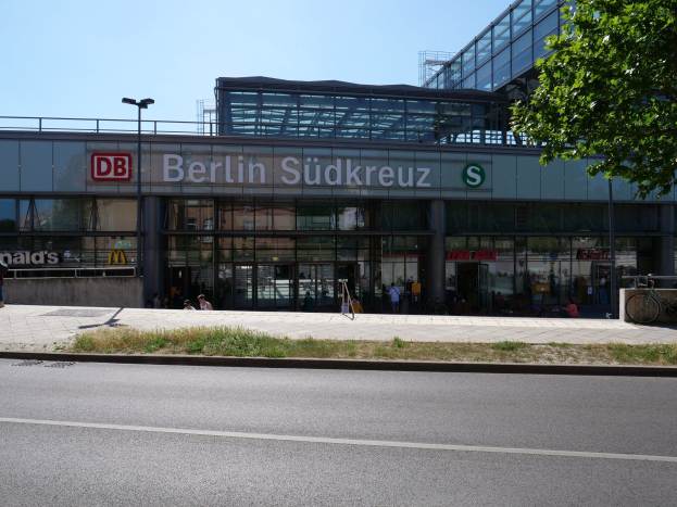Großes Glaswand-Fernbahnhof Berlin Südkreuz mit sichtbarem Text, umgeben von Straßeninfrastruktur, Fahrzeugen, Fußgängern, Fahrrädern, Bäumen und einem klaren Himmel.
