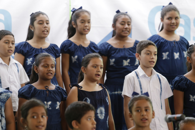 Gruppe von Kindern, die gemeinsam vor einem Mikrofon singen, mit einem Schulchor-Banner im Hintergrund.