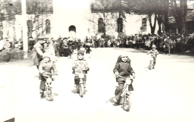 Schwarzes und weißes Bild von Kindern, die Fahrräder eine Straße hinunterfahren, mit einem Gebäude und Bäumen im Hintergrund.