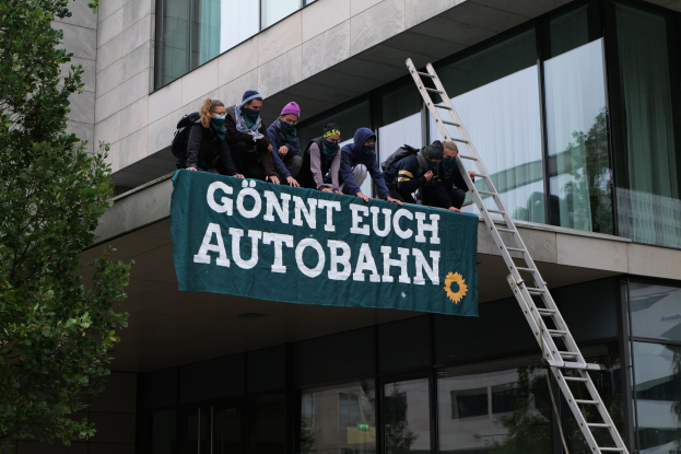 Gruppe von Menschen auf einem Gebäude mit einer Fahne, mit einem Baum links und Glasfenstern im Hintergrund, wahrscheinlich protestierend im Support der deutschen Regierung.