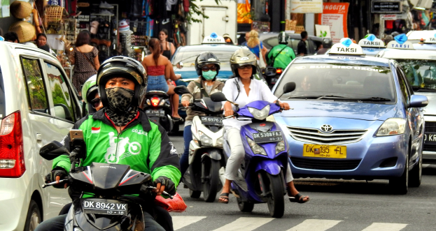 Eine Gruppe von Motorradfahrern fährt auf einer belebten Straße neben Autos und anderen Fahrzeugen, wobei einige Fahrer Helme tragen, und Gebäude, Bäume, Schilder und Geschäfte im Hintergrund zu sehen sind.