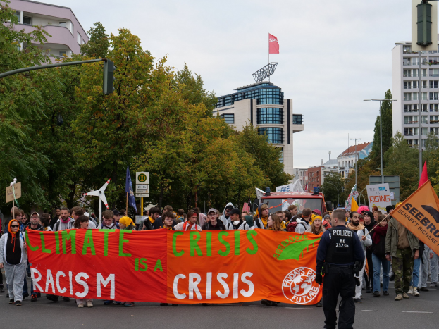Menschen marschieren eine baumgesäumte Straße entlang und halten ein "Klima-Krise ist eine Krise"-Schild mit parkenden Fahrzeugen, Gebäuden und einem klaren Himmel im Hintergrund.