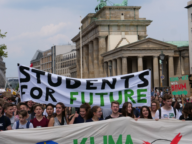 Eine Gruppe von Schülern marschiert in Berlin, eine bunt bemalte "Schüler für die Zukunft"-Fahne tragend, vor einer Kulisse aus Gebäuden, Bäumen und Himmel.