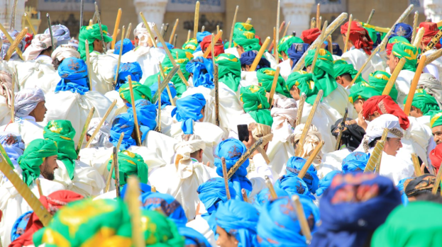 Eine große Gruppe von Menschen in farbenfrohen Kostümen marschiert eine Straße entlang, jeder hält einen Stock in der Hand, im Hintergrund ist ein Gebäude zu sehen.