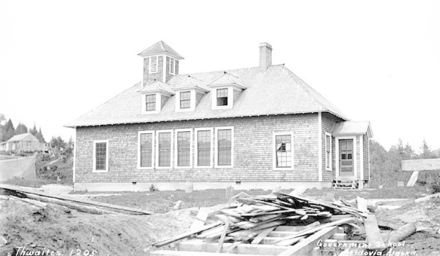 Schwarz-weiß-Fotografie eines zweistöckigen Gebäudes mit mehreren Fenstern und Türen, umgeben von Bäumen und einem klaren Himmel, beschriftet mit "Regierungsschule, Thwaites, 1905" unten.