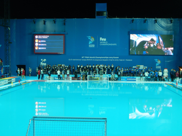 Eine Gruppe von Menschen um einen Swimmingpool mit einem Netz am Boden herumstehend, mit einem Bildschirm im Hintergrund, der "FINA World Championships 2015 Kazan" anzeigt.