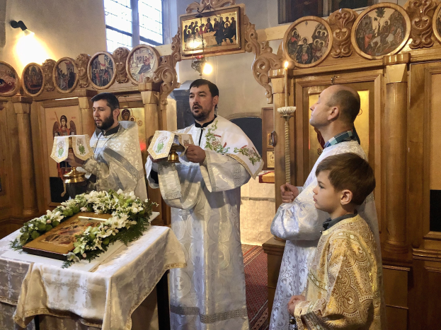 Eine Gruppe von Menschen steht um einen Tisch in einer Kirche mit einem Tuch und einem Blumenstrauß, haltend Bücher, während einer Feier der Ordination der Heiligen Dreifaltigkeit, mit Foto Rahmen, Lampen, einem Fenster und einem hölzernen Objekt im Hintergrund.