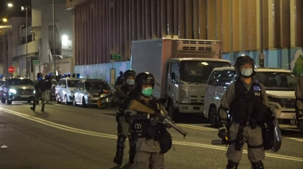 Polizeibeamte in Helmen, Handschuhen und Gesichtsmasken stehen auf einer Straße bei Nacht, einige halten Waffen, mit Fahrzeugen, Laternenmasten, Schildern und Gebäuden im Hintergrund.