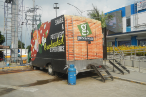 Ein Lastwagen vor einem Gebäude mit einer Tafel, auf der "Teilen Sie eine positiv überladene Erfahrung" steht, mit Stufen, einem Zylinder, Geländern, Strommasten, Laternen, Bäumen und einem bewölkten Himmel.