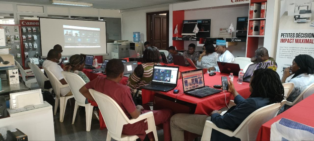Gruppe von Menschen in einem Werkstattsetting mit Laptops, einem Banner, Display-Bildschirm und Deckenbeleuchtung.