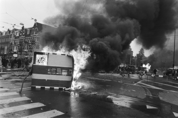 Schwarz-weißes Foto eines brennenden Busses am Straßenrand mit Menschen in der Nähe, umgeben von Pfählen, einem Schild, Bäumen, Gebäuden und einem bewölkten Himmel.