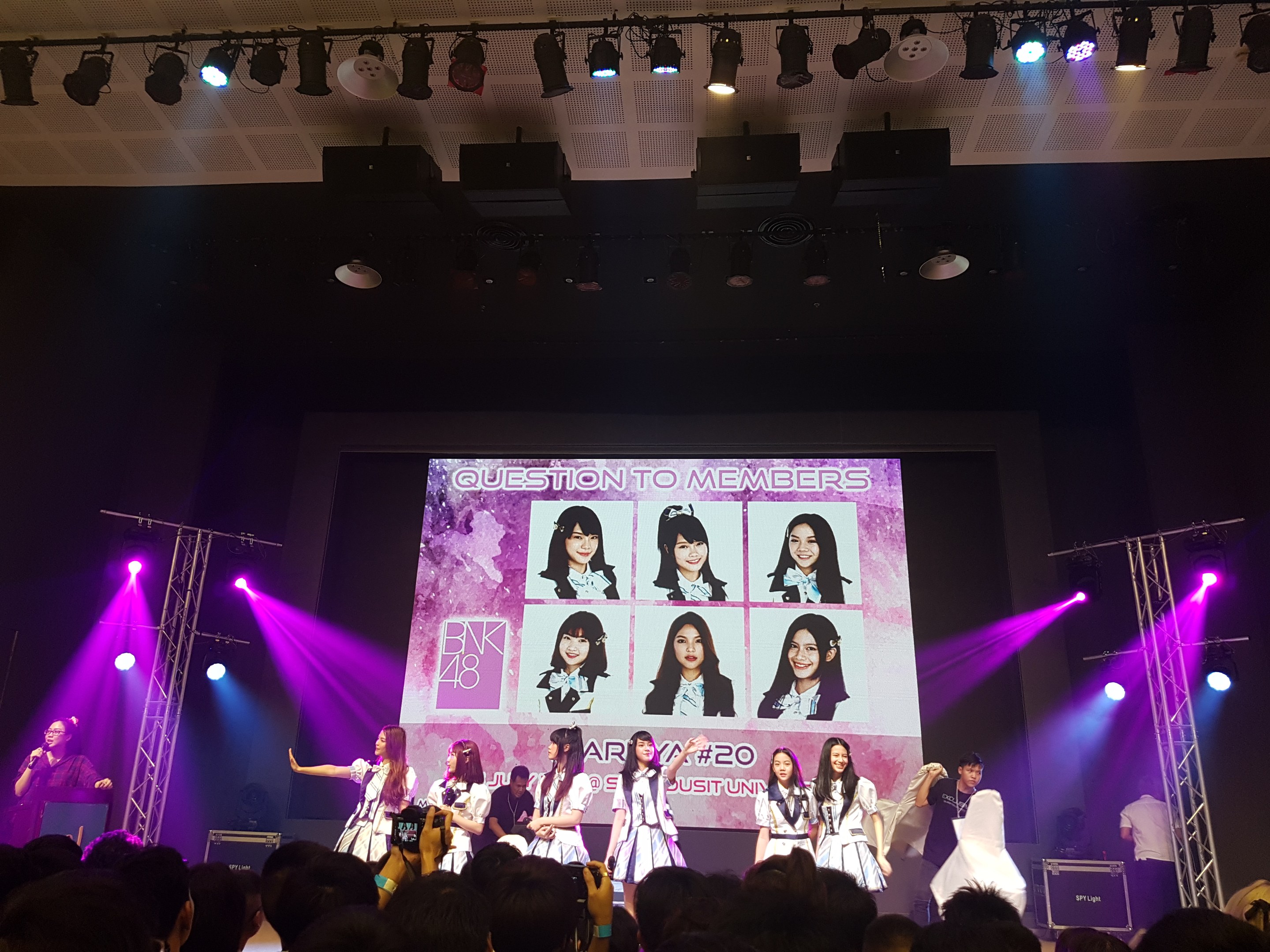 Eine Gruppe von Menschen, die auf einer hell erleuchteten Bühne performen, mit einer Menge darunter, mit einem Sänger mit Mikrofon und einem Musiker, der ein Instrument spielt, und einem großen Bildschirm im Hintergrund, der Bilder von BK48 anzeigt.