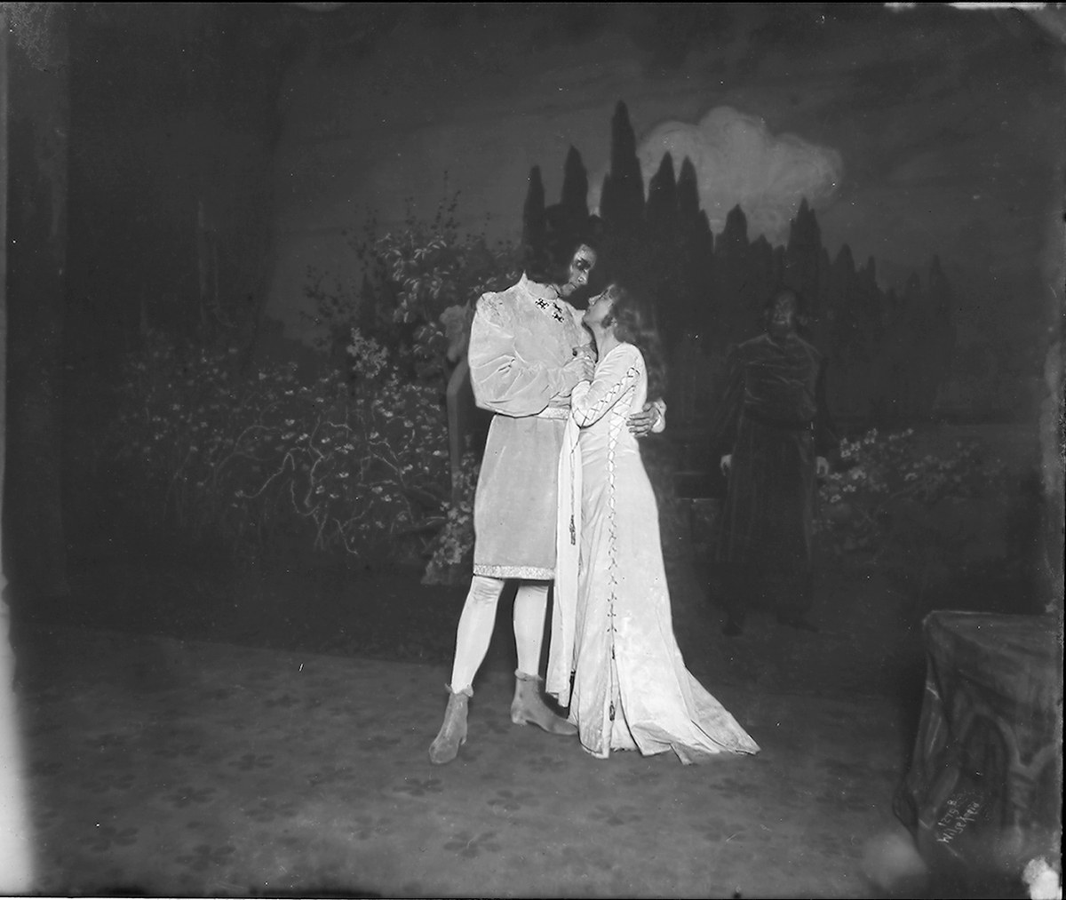 Schwarzes und weißes Foto eines Mannes und einer Frau, die sich auf einer Bühne umarmen und küssen, mit einem Publikum, Pflanzen und Bäumen im Hintergrund.