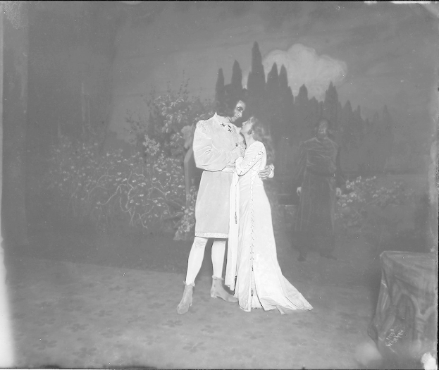 Schwarzes und weißes Foto eines Mannes und einer Frau, die sich auf einer Bühne umarmen und küssen, mit einem Publikum, Pflanzen und Bäumen im Hintergrund.