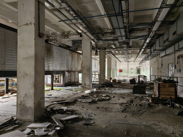 Ein verlassenes Gebäude mit Schutt, einschließlich gebrochener Holzteile und Metall, auf dem Boden verstreut, Säulen, Fenster mit Jalousien, eine Wand im Hintergrund und eine Decke mit Rohren oben.
