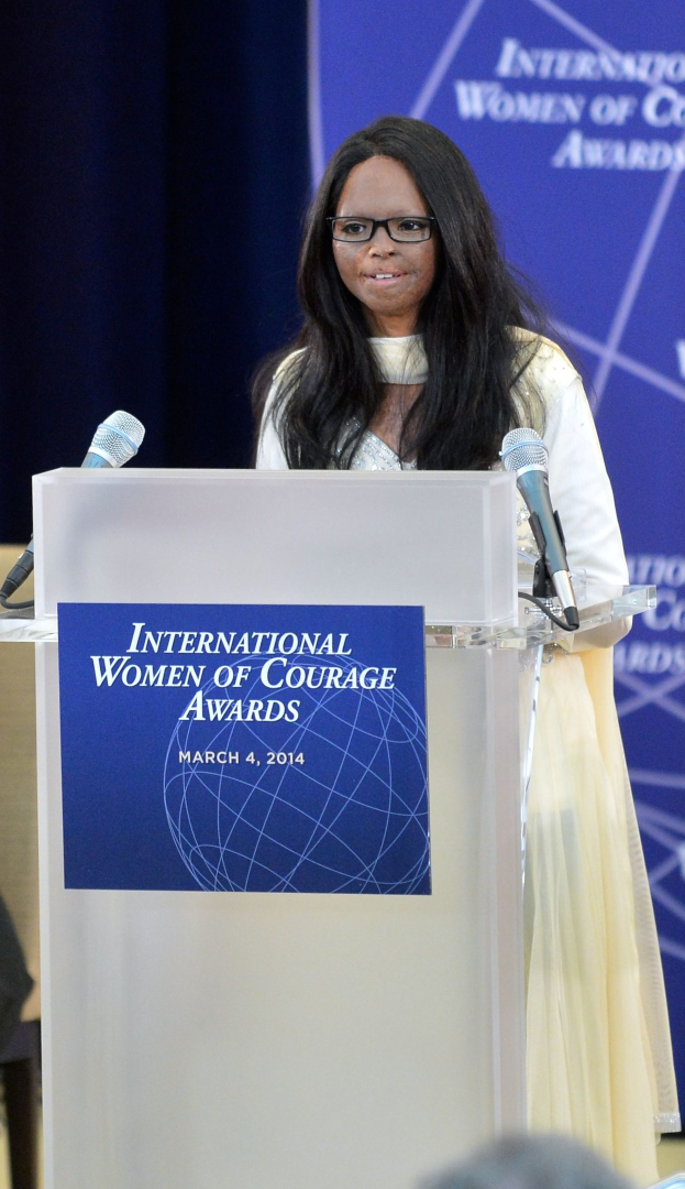 Eine Frau In Einem Weißen Kleid Und Brille Steht An Einem Podium Mit Zwei Mikrofonen Und Einer Tafel, Redet Vor Einer Menge Hinter Einer Blauen Flagge Mit Der Aufschrift "International Women Of Courage Awards 2014".