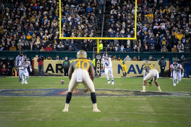 Fußballspieler in Sportbekleidung und Helmen konkurrieren auf einem Feld mit Zuschauern im Hintergrund, Stadionstrukturen und sichtbare Beschilderung.