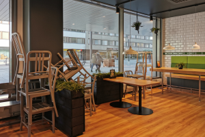 Ein Restaurant in Berlin, Deutschland, mit Holztischen und -stühlen in der Nähe eines großen Fensters, das Menschen auf der Straße, Gebäude und den Himmel zeigt, umgeben von Pflanzen und Lichtern.