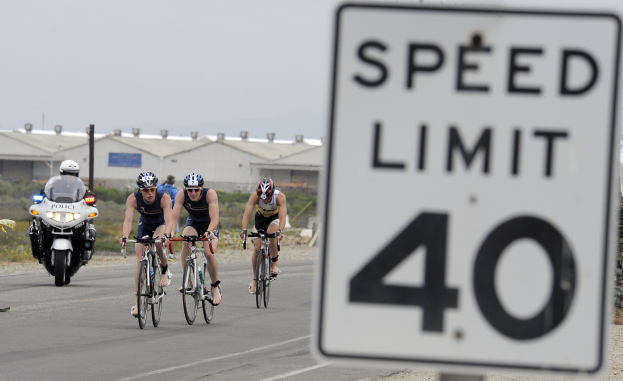 Eine Gruppe von Radfahrern mit Helmen fährt auf einer Straße neben einem Tempolimitschild vorbei, mit Gebäuden, Masten, Pflanzen, Bäumen und einem klaren blauen Himmel im Hintergrund.