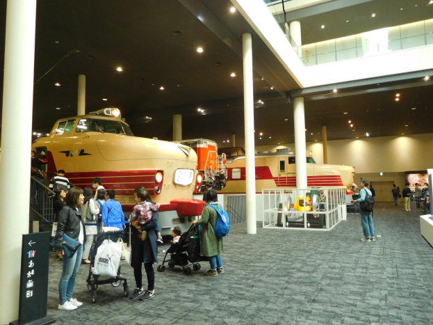 Gruppe von Menschen bei der Betrachtung von Oldtimer-Zügen in einem Museum mit Säulen, Geländern und Deckenbeleuchtung, einige mit Taschen und Kinderwagen und ein Textschild links sichtbar.