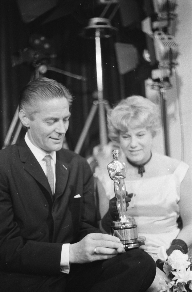 Ein Mann in einem schwarzen Blazer, weißem Hemd und Krawatte hält eine Oscar-Statue in der Hand und sitzt neben einer Frau, die einen Blumenstrauß hält, beide in formeller Kleidung, mit ein paar Lichtern im schwarzen und weißen Hintergrund.