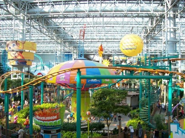 Indoor-Wasserpark mit einer Achterbahn, umgeben von Menschen, Bäumen, Pflanzen und beleuchteten Deckenstangen.