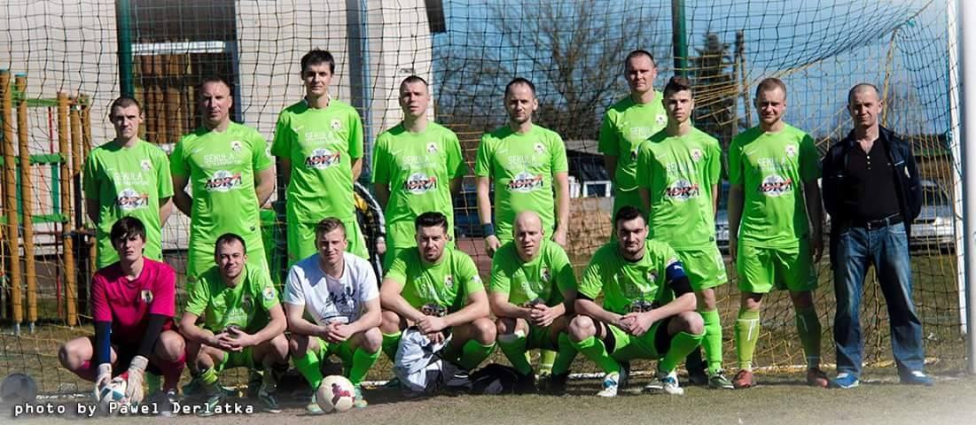 Eine Gruppe von Männern in grünen T-Shirts steht oder hockt auf einem Fußballfeld mit einem Ball davor, in der Nähe eines Torpfostens, mit Bäumen, Gebäuden und Fahrzeugen im Hintergrund.