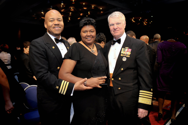 Zwei Männer in schwarzen Blazern und weißen Hemden stehen neben einer Frau in einem schwarzen Kleid, die eine Trophäe hält, auf einem roten Teppich, während andere sitzen und stehen und der Hintergrund von beleuchteten Decken erhellt wird.