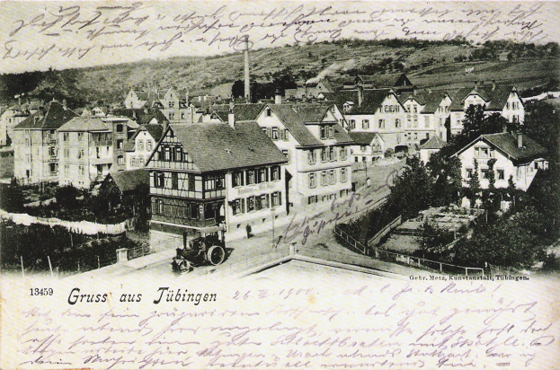 Eine alte Postkarte zeigt eine Stadtansicht mit Gebäuden, Bäumen und einem Fahrzeug auf der Straße, mit Text oben und unten.