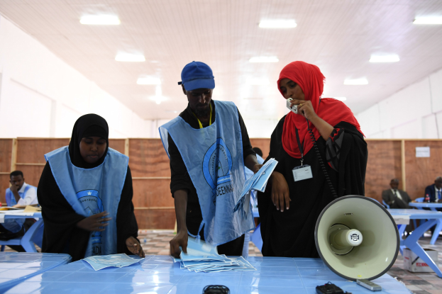 Eine Gruppe von Menschen steht um einen Tisch mit einem Megafon und Papieren, während andere im Hintergrund eines Wahllokals mit beleuchteter Decke sitzen und stehen.