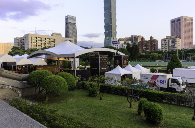 Eine Gruppe von Zelten in einem Park mit Grünflächen, Menschen, Fahrzeugen, Gebäuden und einem klaren Himmel, wahrscheinlich beim Hong Kong International Film Festival.