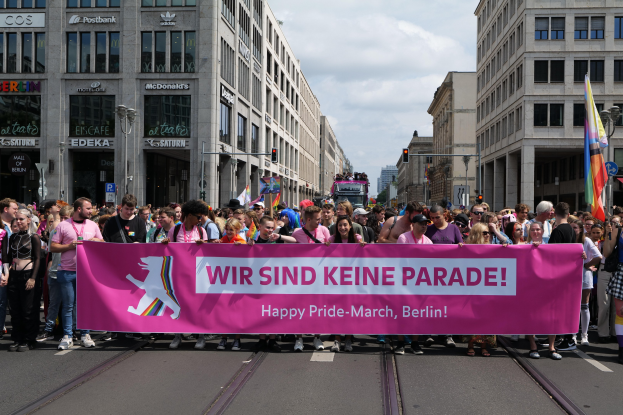 Eine Gruppe von Menschen marschiert auf einer Berliner Straße und hält ein pinkes "Happy Pride March"-Schild, während Gebäude, Laternenpfähle und Verkehrszeichen die Straße säumen.