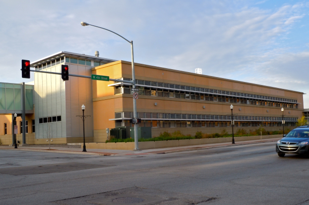Ein Auto fährt auf einer Straße neben einem hohen Gebäude mit Fenstern, Laternen, Verkehrszeichen, Schildern, Pflanzen, Bäumen und einem bewölktem Himmel im Hintergrund.