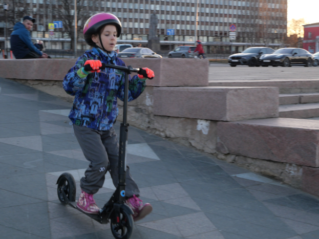 Ein junger Junge in einem Helm und Handschuhen fährt auf einem Roller eine Treppe hinunter, mit Autos, Menschen, Bäumen, Pfosten, Brettern, Gebäuden und einem klaren blauen Himmel im Hintergrund.
