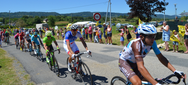 Eine Gruppe von Radfahrern fährt eine Straße entlang, während Zuschauer am Straßenrand stehen, einige tragen Helme und Schutzbrillen oder halten Fahnen, mit Bäumen, Schildern, Fahrzeugen, Zelten, Gebäuden, Hügeln und einem klaren blauen Himmel im Hintergrund.