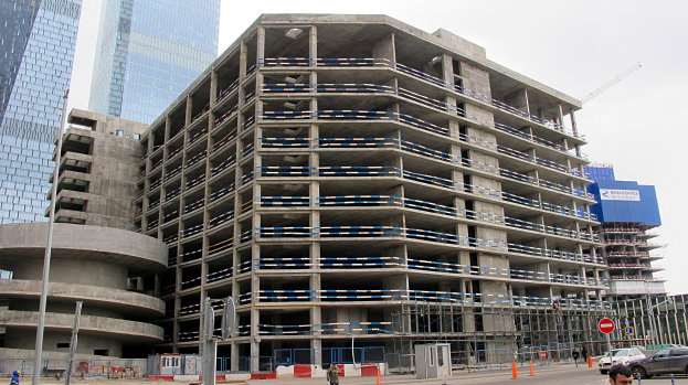 Ein großes Bauwerk im Bau in einer städtischen Umgebung, umgeben von Wolkenkratzern, mit Fahrzeugen, Fußgängern, Verkehrsinfrastruktur und Baumaschinen unter einem klaren Himmel.