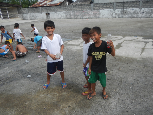 Gruppe junger Jungs auf einem Parkplatz, einige halten Flaschen, mit Häusern, Bäumen, Pfählen und einem klaren blauen Himmel im Hintergrund.