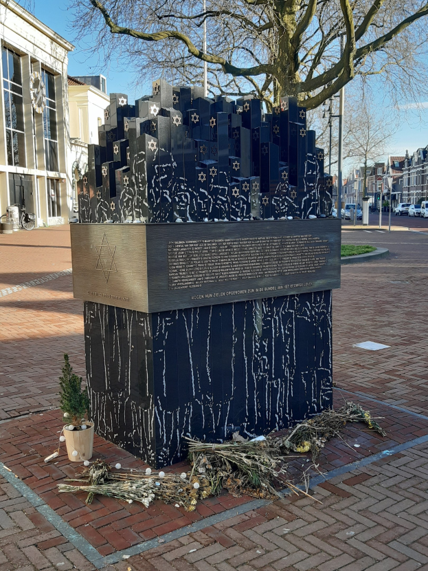 Denkmal in einem Stadtplatz, das an Holocaust-Opfer erinnert, mit einem Baum davor, einem Topfpflanzen auf dem Boden, verstreuten trockenen Blättern und Gebäuden, Fahrzeugen, Masten und Bäumen im Hintergrund.