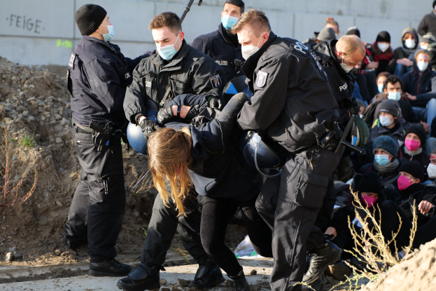 Gruppe maskierter Personen in Mützen, einige bewaffnet, die eine Frau umgeben, die von der Polizei eskortiert wird, mit städtischer Vegetation und einer protestbezogenen Wandnachricht im Hintergrund.