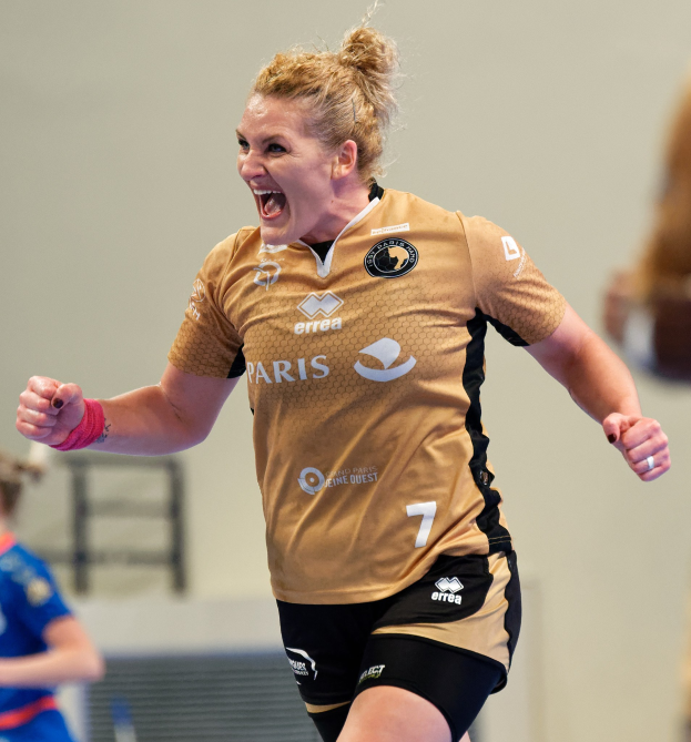 Eine Frau in braunem T-Shirt und schwarzen Shorts rennt auf einem Futsalplatz, feiert mit triumphaler Miene, während zwei andere Frauen im Hintergrund zu sehen sind.